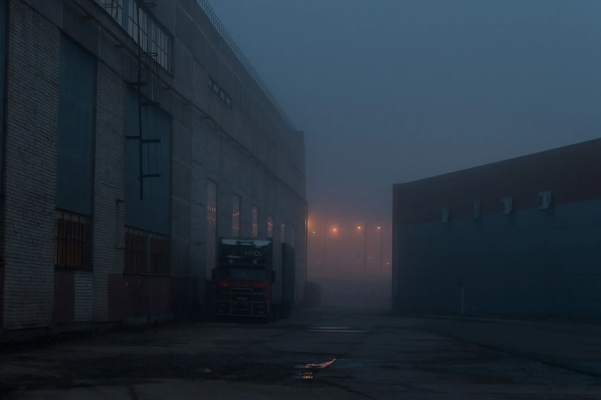 A works yard in fog at dusk, one truck parked against the wall of a long brick building